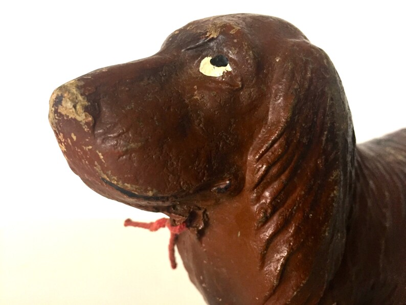 May include: Close-up of a brown ceramic dog figurine with a white eye. The dog has a red ribbon tied around its neck.