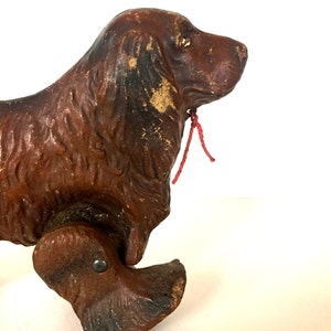 May include: A vintage wooden toy dog with brown paint and a red string attached to its neck. The dog has a long snout and is on wheels.