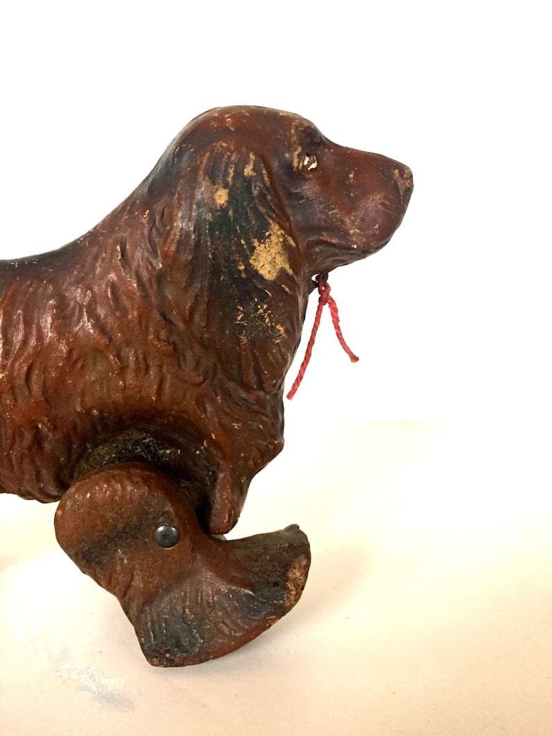 May include: A vintage wooden toy dog with brown paint and a red string attached to its neck. The dog has a long snout and is on wheels.