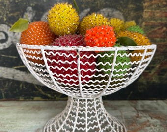 May include: A white wire fruit bowl filled with colorful, beaded fruit. The bowl has a pedestal base and a decorative, openwork design. The fruit includes yellow, orange, red, and green beaded spheres, with a few green leaves.