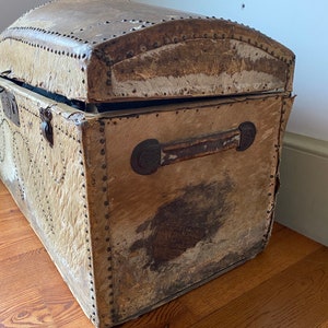 May include: A vintage, brown leather-covered trunk with metal accents and studded details. The trunk is open, revealing the interior lining. The trunk is sitting on a wooden floor.