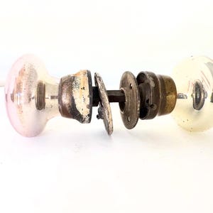 May include: An antique door knob set with two glass knobs, one pink and one yellow, connected by a metal rod and mounting hardware. The hardware shows signs of age and wear, with a distressed appearance.