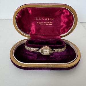 May include: A gold-colored wristwatch with a white face and black hands, in a purple velvet lined oval box with a gold trim. The box lid has the words "BENRUS OFFICIAL WATCH OF FAMOUS AIRLINES" printed in gold.