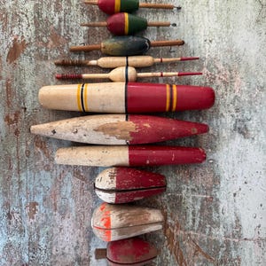 Puede incluir: Una colección de flotadores de pesca de madera vintage en varias formas y tamaños. Los flotadores están pintados en una variedad de colores, incluyendo rojo, blanco, verde y amarillo. Algunos tienen rayas u otros elementos decorativos. Dispuestos verticalmente sobre un fondo de madera envejecida.