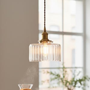 Op de afbeelding: Een heldere glazen hanglamp met een geribbelde kap hangt boven een aanrecht. De lamp heeft een messingkleurige basis en een zichtbare gloeilamp. Een koffiezetapparaat, een mok en gebak staan op het aanrecht.