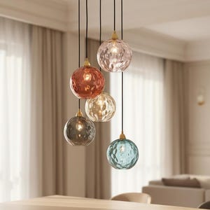 May include: A modern chandelier with five textured glass globes in various colors: pink, red, gold, gray, and blue. The globes hang from black cords attached to a gold-colored circular ceiling plate. The fixture is suspended above a wooden table.