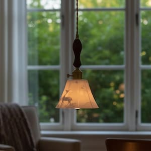 Peut inclure: Une lampe suspendue avec un abat-jour crème orné d'une scène de cerf. La lampe a un accent en bois brun foncé et un raccord en laiton. Elle est suspendue au-dessus d'une table en bois avec un livre et une tasse à thé.