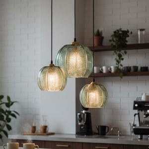 May include: Three pendant lights with a textured, green glass shade hang above a kitchen counter. The shades have a ribbed pattern and are illuminated from within. The kitchen has a coffee machine, mugs, and a brick wall.