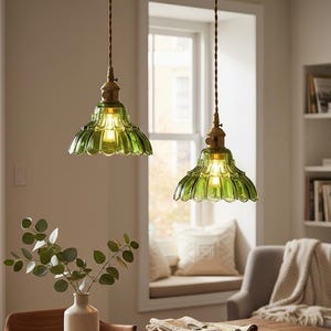Może przedstawiać: Dwie zielone szklane lampy wiszące z muszelkowym wzorem wiszą nad drewnianym stołem jadalnym. Lampy mają mosiężne oprawy i są podświetlane. Wazon z zielenią i stos białych talerzy znajdują się na stole.
