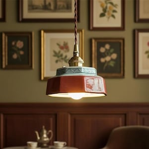 Puede incluir: Lámpara colgante con pantalla octogonal roja y banda decorativa azul y blanca. La lámpara tiene una parte superior de color latón y un cable trenzado marrón. El fondo presenta grabados botánicos enmarcados y una mesa de té.
