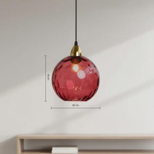 May include: A red glass pendant light with a textured surface, 7 inches in diameter and 7 inches high. The light fixture has a gold-colored metal base and a black cord. The light is hanging in front of a white wall.