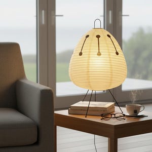 Op de afbeelding: Crèmekleurige papieren lamp met een zwart metalen driepoot. De lamp heeft een druppelvorm met donkerbruine accenten. De lamp staat op een houten tafel naast een kopje thee en een bril.