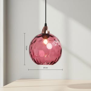 May include: A pink glass pendant light with a textured surface. The light fixture is 18 cm high and 20 cm wide. It has a copper-colored base and a black cord. The light is hanging in front of a light-colored wall.