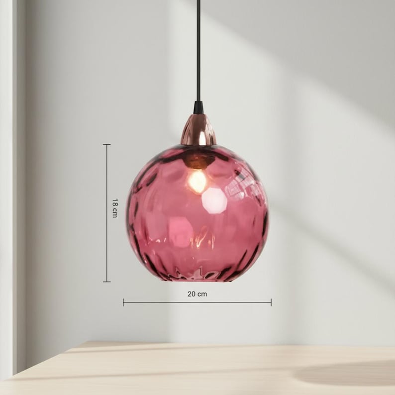 May include: A pink glass pendant light with a textured surface. The light fixture is 18 cm high and 20 cm wide. It has a copper-colored base and a black cord. The light is hanging in front of a light-colored wall.