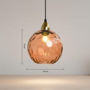 May include: A pendant light with a textured, spherical, amber-colored glass shade, 18 cm in height and 20 cm in diameter. The light fixture has a gold-colored metal cap and a black cord. The light is turned on, illuminating the interior.