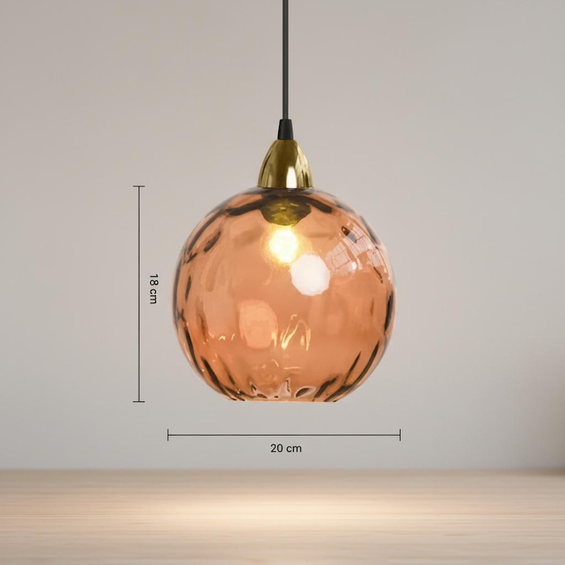 May include: A pendant light with a textured, spherical, amber-colored glass shade, 18 cm in height and 20 cm in diameter. The light fixture has a gold-colored metal cap and a black cord. The light is turned on, illuminating the interior.