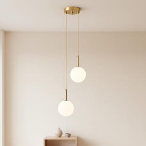Könnte beinhalten: Zwei Pendelleuchten mit kugelförmigen, weißen Glasschirmen hängen an einer goldfarbenen Deckenplatte. Die Leuchten haben ein minimalistisches Design mit goldfarbenen Stäben und Schnüren, wodurch eine moderne Ästhetik entsteht. Der Hintergrund ist eine neutrale, hellfarbige Wand.