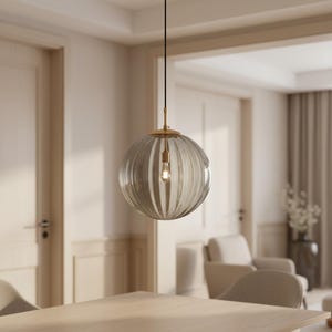 May include: A clear, ribbed glass pendant light with a gold-colored metal fixture. The light bulb is visible inside the globe-shaped shade. The light hangs from a black cord in a room with neutral tones.