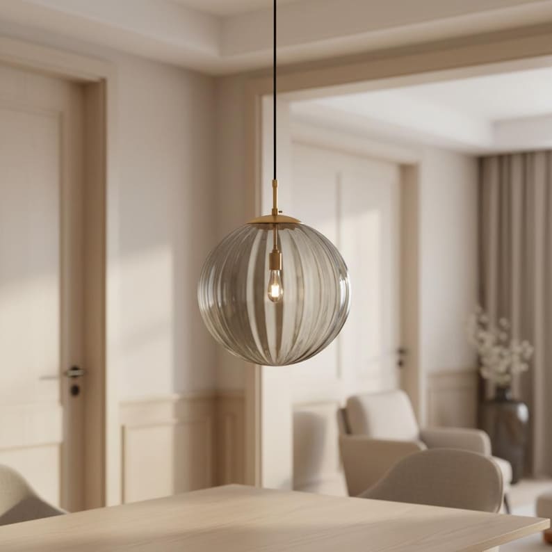 May include: A clear, ribbed glass pendant light with a gold-colored metal fixture. The light bulb is visible inside the globe-shaped shade. The light hangs from a black cord in a room with neutral tones.