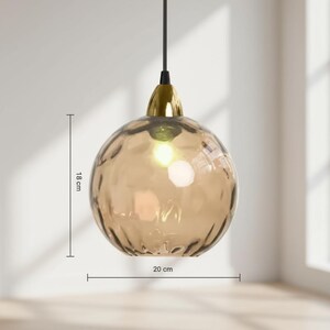 May include: A pendant light fixture with a textured, amber-colored glass globe, approximately 7 inches in diameter and 7 inches tall. The fixture has a gold-colored metal cap and a black cord. The light is turned on, illuminating the interior.