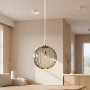 May include: A clear, ribbed glass pendant light with a brass-colored fixture and a visible lightbulb. The light hangs from a black cord, creating a modern and elegant aesthetic. The spherical shape and neutral tones make it suitable for various interior styles.
