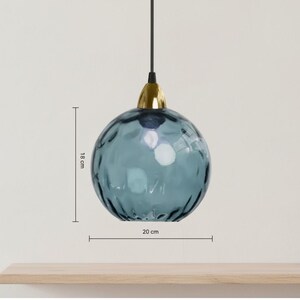 May include: A blue glass pendant light with a textured surface, 7 inches in diameter and 7 inches tall. It has a gold-colored metal cap and a black cord. The light hangs from the ceiling, showcasing a modern design.