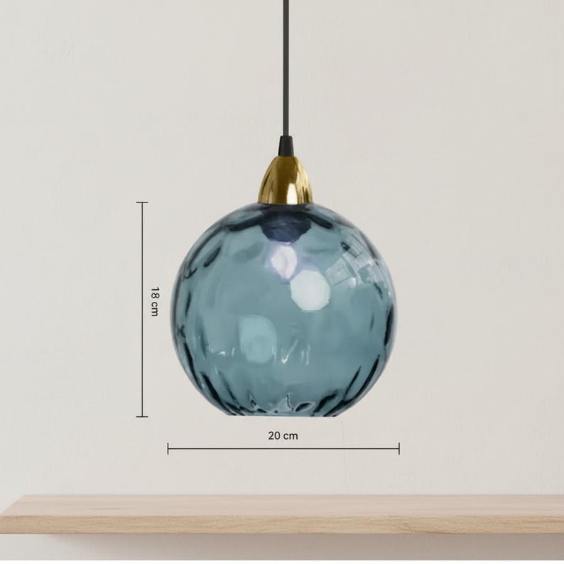 May include: A blue glass pendant light with a textured surface, 7 inches in diameter and 7 inches tall. It has a gold-colored metal cap and a black cord. The light hangs from the ceiling, showcasing a modern design.