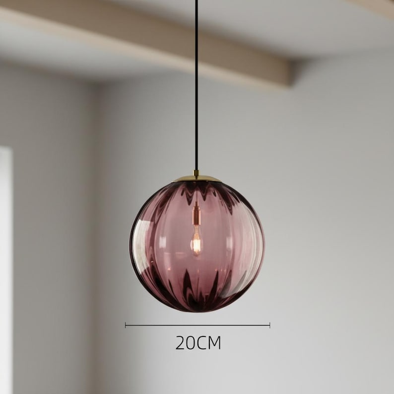May include: A pendant light fixture with a spherical, ribbed, purple glass shade. The shade is 20cm in diameter and hangs from a black cord with a gold-colored metal cap. The light bulb is visible inside the shade.