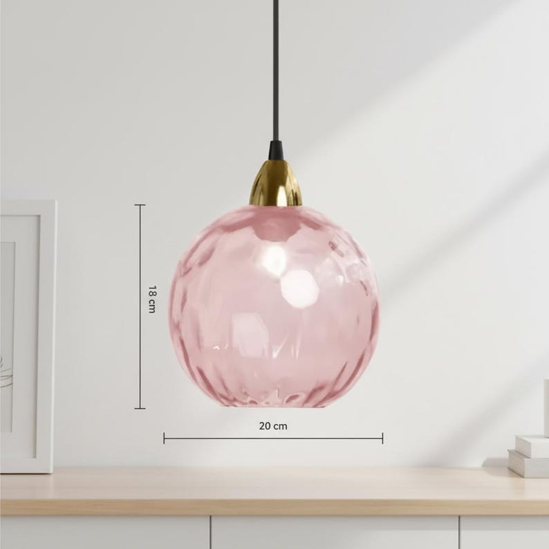 May include: A pink glass pendant light with a textured surface, 7 inches in height and 8 inches in diameter. The light fixture has a gold-colored base and a black cord. The light is hanging from the ceiling.