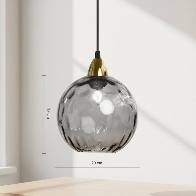 May include: A gray glass pendant light with a textured surface, 18 cm high and 20 cm wide. It has a gold-colored metal base and a black cord. The light is spherical and hangs from the ceiling, suitable for home decor.