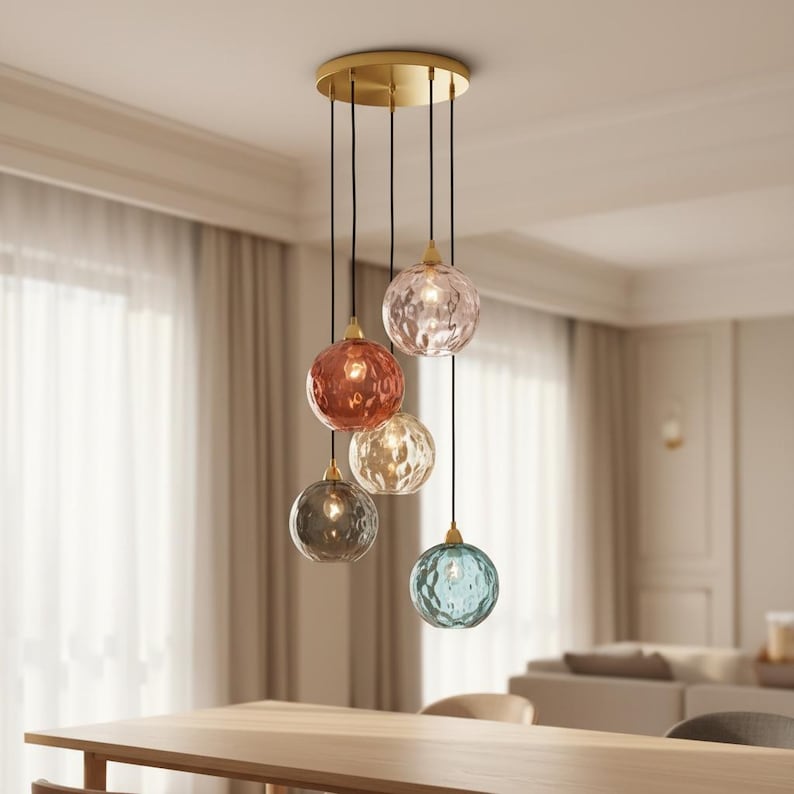 May include: A modern chandelier with five textured glass globes in various colors: pink, red, gold, gray, and blue. The globes hang from black cords attached to a gold-colored circular ceiling plate. The fixture is suspended above a wooden table.