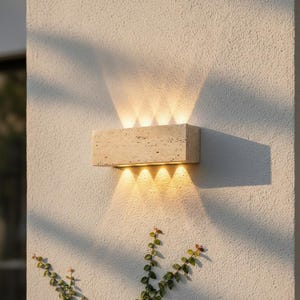 Pode incluir: Uma luminária de parede retangular para exterior feita de pedra bege. A luz emana da parte superior e inferior, criando um brilho quente. A luminária é montada em uma parede branca texturizada, com videiras verdes em primeiro plano.