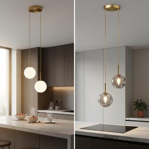 Puede incluir: Dos lámparas colgantes con pantallas esféricas de cristal. Las luces tienen un acabado dorado y cuelgan del techo. Una luz tiene una pantalla blanca opaca, y la otra tiene una pantalla transparente, teñida de gris. Las luces están suspendidas sobre una isla de cocina.