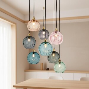 May include: A cluster of eight pendant lights with textured, colored glass globes. The globes are in shades of pink, blue, gray, and green, each with a gold-colored cap. The lights hang from black cords, creating a modern lighting fixture.