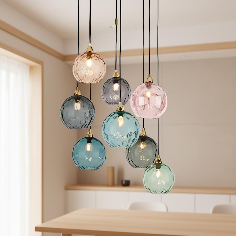 May include: A cluster of eight pendant lights with textured, colored glass globes. The globes are in shades of pink, blue, gray, and green, each with a gold-colored cap. The lights hang from black cords, creating a modern lighting fixture.