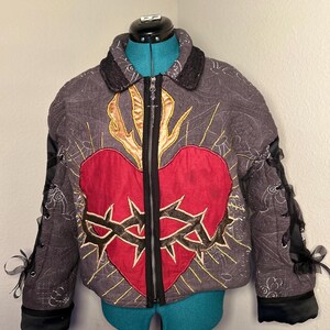 May include: A quilted jacket with a central red heart design encircled by a gold-stitched crown of thorns. The jacket features a black zipper, black lace-up sleeves, and a decorative collar. The background is a muted gray with gold stitching.