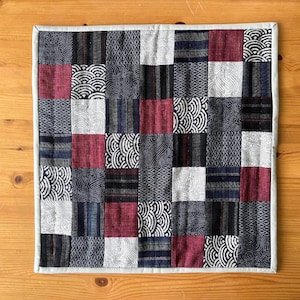 May include: Square patchwork quilt with a variety of patterned fabrics in shades of navy, burgundy, white, and gray. The quilt is bordered with a light gray fabric and is displayed on a wooden surface.