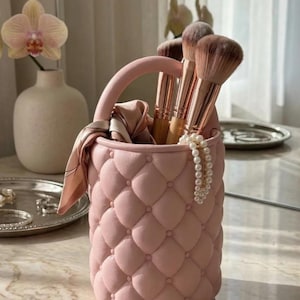 May include: A pink quilted makeup brush holder with a handle, containing makeup brushes. A silk scarf and a pearl necklace are draped over the side. The holder is on a marble surface, with a vase and a tray in the background.