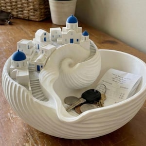 Könnte beinhalten: Dekorative Schale aus weißer Keramik mit Muscheldesign. Verfügt über ein Miniaturdorf von Santorin mit blau gewölbten Dächern. Enthält Schlüssel, Münzen und eine Quittung.