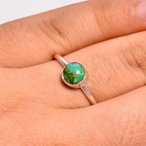 Puede incluir: Un anillo de plata con una piedra preciosa de turquesa redonda tallada en cabujón, con patrones de matriz en tonos verdes y marrones. La piedra está engastada en un bisel de plata pulida sobre una banda de plata fina y lisa.