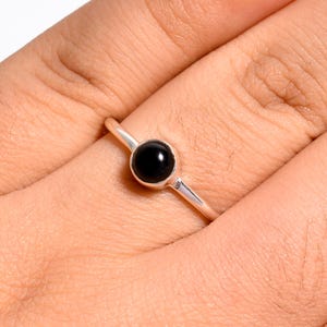 May include: A silver ring featuring a single, smooth, round black stone set in a simple metal bezel. The band is thin and polished, showcasing a minimalist design that highlights the contrast between the dark gemstone and the bright silver metal.