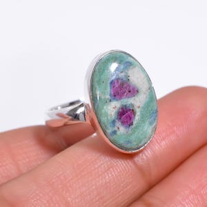 May include: A silver ring featuring a large, oval-shaped cabochon gemstone. The stone displays a mottled pattern of light green and white minerals with two distinct, vibrant magenta-colored inclusions embedded within the surface of the polished stone.