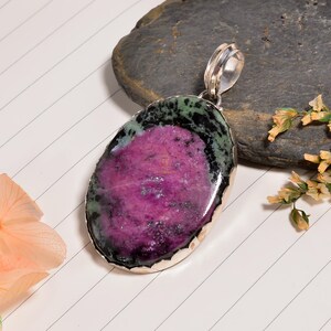 Puede incluir: Colgante ovalado con una piedra magenta en el centro, bordeada por verde oscuro y negro, engastada en un marco plateado. El colgante cuelga de una fianza plateada. Fondo blanco con líneas diagonales.