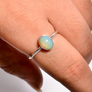 May include: A silver ring featuring a smooth, oval-shaped opal gemstone with iridescent flashes of green, orange, and yellow. The stone is set in a simple, polished silver band that encircles the finger, showcasing a clean and reflective metal finish.