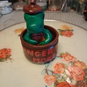 Peut inclure: Une petite figurine de dinosaure vert portant un béret marron, assise dans un pot cylindrique marron. Le pot porte l'inscription "DANGER MORD" en lettres rouges sur le devant. La figurine présente une finition lisse et brillante.