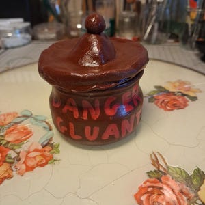 Peut inclure: Un petit pot en argile sculpté à la main de couleur brune avec un couvercle assorti doté d'un bouton sphérique. L'extérieur du pot est peint avec le texte rouge "DANGER GLUANT" dans un style manuscrit gras, indiquant une étiquette d'avertissement.