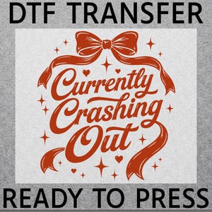 May include: A DTF transfer with the text "DTF TRANSFER" at the top and "READY TO PRESS" at the bottom. The design features the words "Currently Crashing Out" in a decorative font, with a bow, ribbons, hearts, and stars.