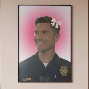 May include: Framed print featuring a smiling man in a police uniform with a pink bow in his hair. The background is a gradient of pink and white. The print is displayed on a light pink wall.