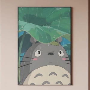 Op de afbeelding: Een ingelijste illustratie van een grijze, pluizige figuur uit de animatiefilm "My Neighbor Totoro", schuilend onder een groot groen blad in de regen. Het kunstwerk heeft een zwarte lijst en een lichtbeige achtergrond.