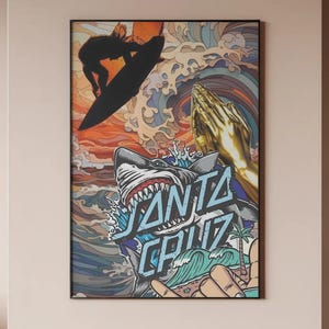Op de afbeelding: Een rechthoekige wandkunstprint in een zwarte lijst met een collage van een surfer-silhouet, gouden biddende handen, een haaienkop en oceaangolven. De tekst SANTA CRUZ staat in blauwe letters prominent over de afbeelding heen.